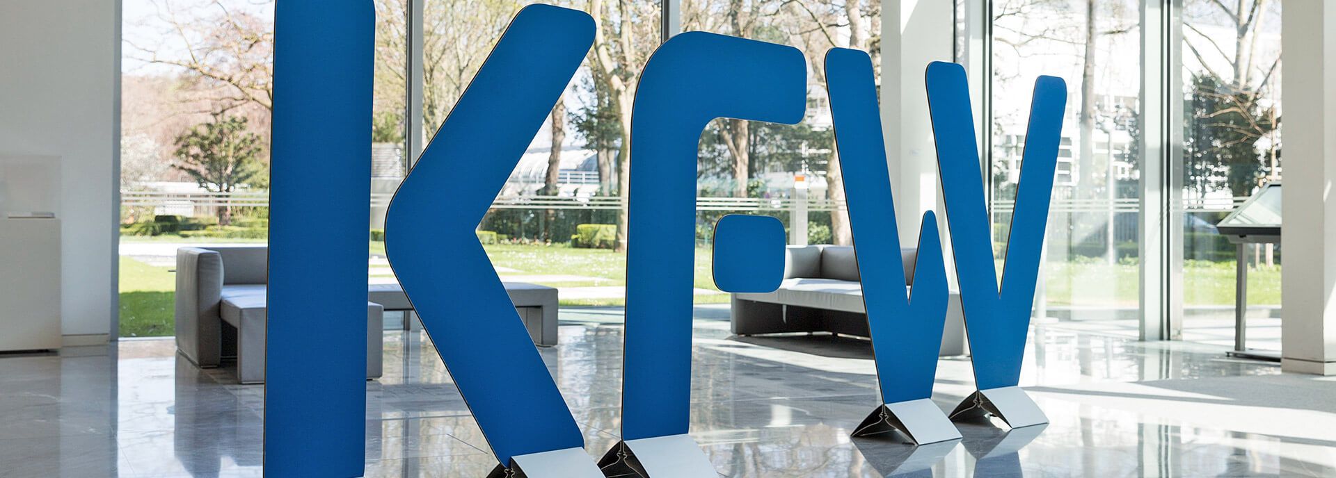 Große, blaue KFW-Buchstaben in einer modernen Lobby mit Glasfront und Sofas.