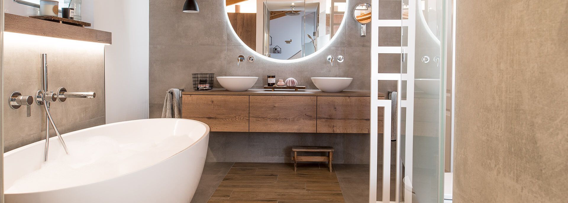 Freistehende Badewanne, Doppelwaschtisch aus Holz, runder Leuchtspiegel und Wände in Betonoptik.