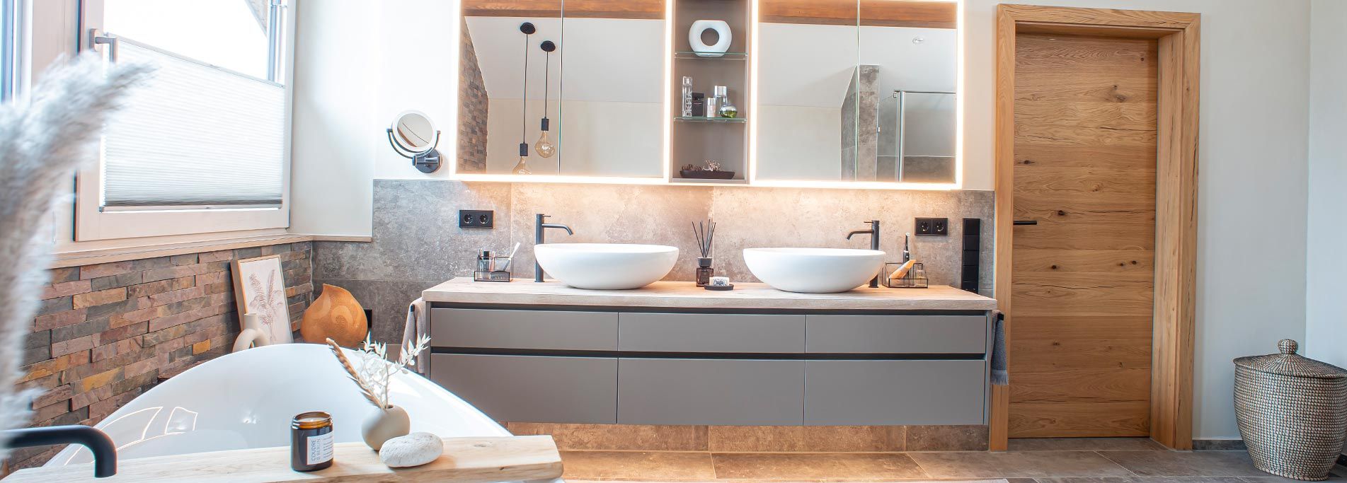 Doppelwaschtisch mit Aufsatzwaschbecken, schwarzen Armaturen, beleuchtetem Spiegelschrank und Wandverkleidung in Steinoptik.