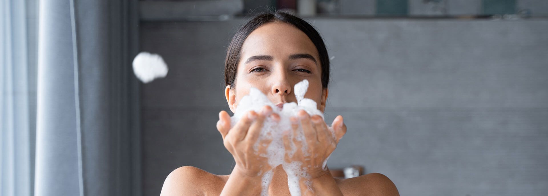 Wir sorgen für mehr Freude im Badezimmer Junge Frau bläst Badewannen-Schaum aus den Händen, lächelt glücklich.